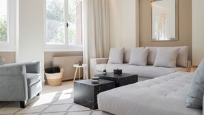 A modern living room with a grey sofa, a white sofa, and a grey armchair. There are two marble coffee tables, a small side table, and a wicker basket. A large window with curtains and a radiator are visible. - Elaia House Glyfada (Glyfada)