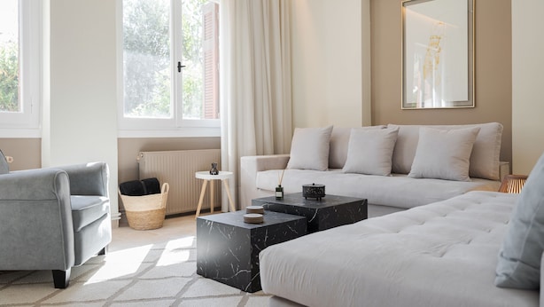 A modern living room with a grey sofa, a white sofa, and a grey armchair. There are two marble coffee tables, a small side table, and a wicker basket. A large window with curtains and a radiator are visible. - Elaia House Glyfada (Glyfada)