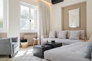 Ein modernes Wohnzimmer mit einem grauen Sofa, einem weißen Sofa und einem grauen Sessel. Zwei Marmortische, ein kleiner Beistelltisch und ein Korbwäschekorb sind vorhanden. Ein großes Fenster mit Vorhängen und eine Heizung sind sichtbar.
