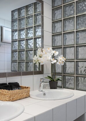 A bathroom with a white sink, a mirror, and a window with a marble pattern. There is a basket with towels and a potted plant on the sink. - Elaia House Glyfada (Glyfada)
