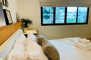 Ein Hotelzimmer mit Bett, Nachttisch mit Lampe, einem Fenster mit Jalousien und Blick auf Gebäude.