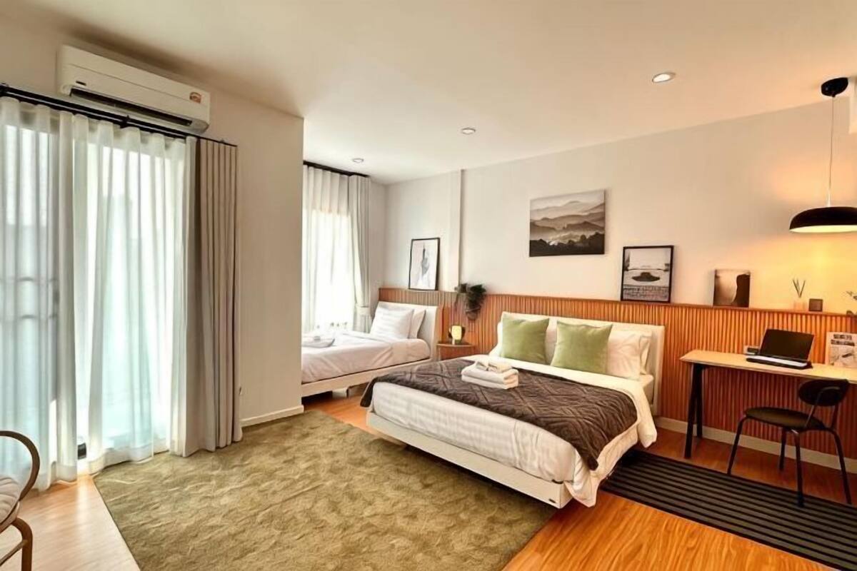 Quarto de hotel com cama, escrivaninha, cadeira, janela com cortinas e luminária na parede.