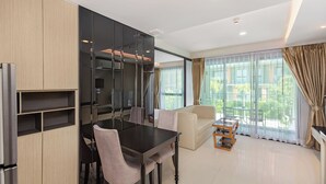 A modern living room with a dining area, large windows, and a view of greenery outside. - 1-bedroom Condo Mida Grand Surin Phuket (Choeng Thale)