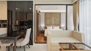 A modern hotel room with a dining area, a sofa, a glass coffee table, a bed with a headboard, and a view of the city through large windows. - 1-bedroom Condo Mida Grand Surin Phuket (Choeng Thale)