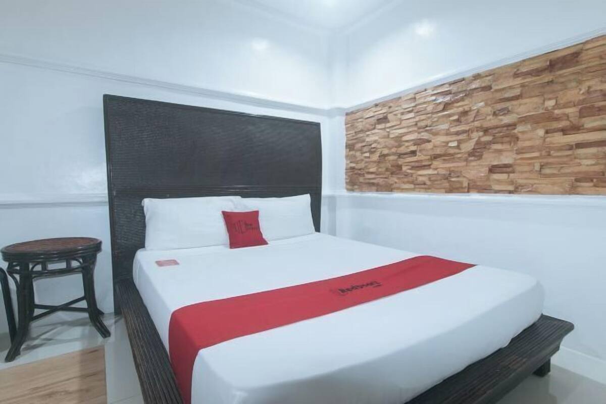 A hotel room with a bed, a small wooden stool, a stone-textured wall, and a red bed runner.