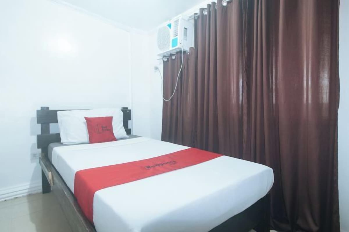 A hotel room with a bed, a red throw, white bedding, a window with curtains, and an air conditioning unit.