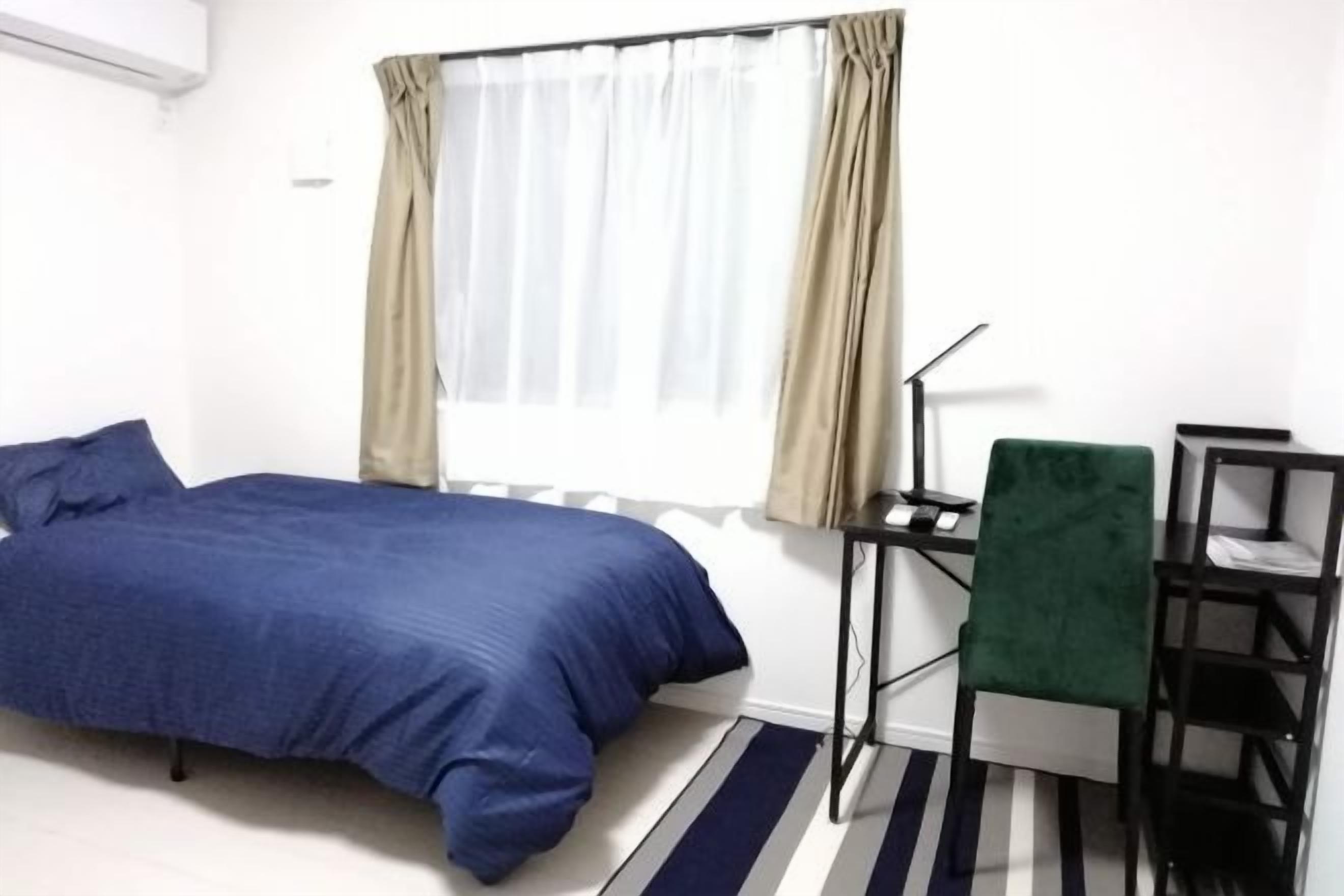 Uma cama de solteiro com colcha azul, uma escrivaninha com cadeira, uma luminária e uma janela com cortinas.
