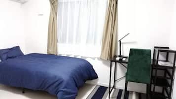 Uma cama de solteiro com colcha azul, uma escrivaninha com cadeira, uma luminária e uma janela com cortinas.