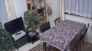 A dining area with a table covered by a floral tablecloth, surrounded by chairs. A wooden bookshelf with various items is against the wall, and a potted plant is on a small table beside the bookshelf.