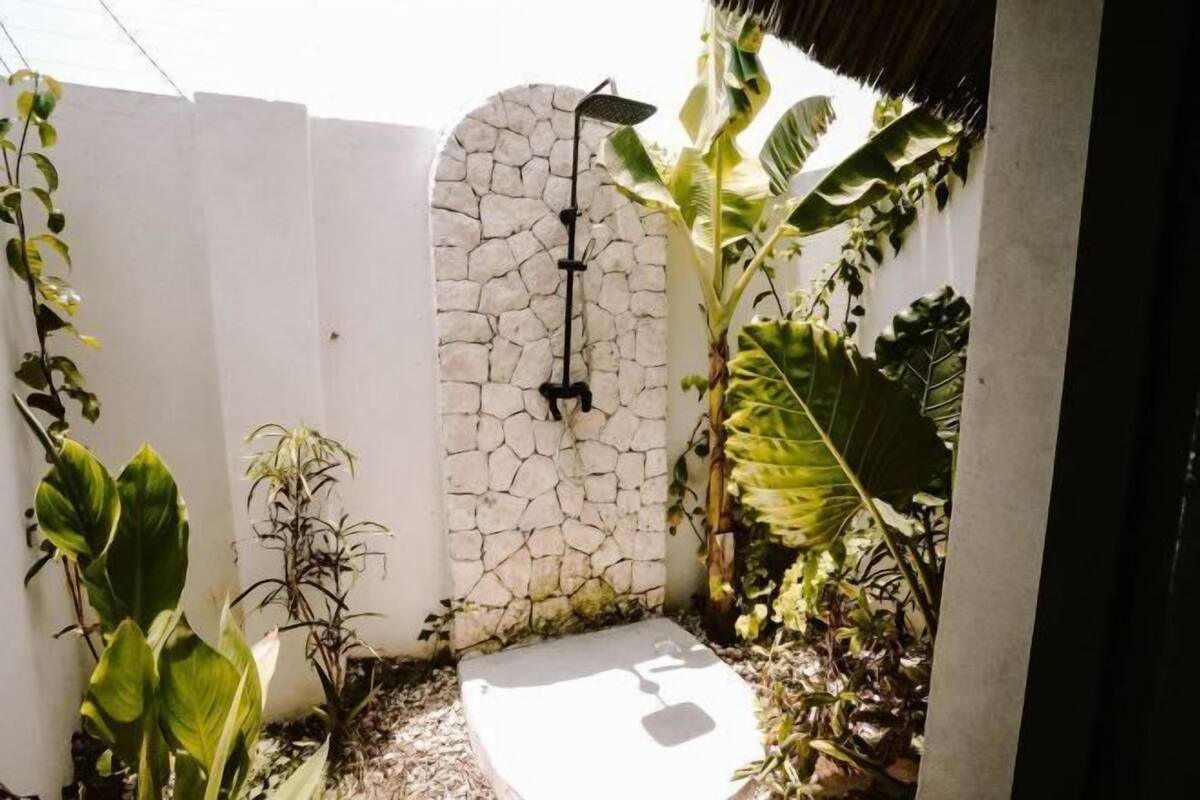 A small, enclosed outdoor shower area with a stone wall, a white fence, and lush tropical plants.