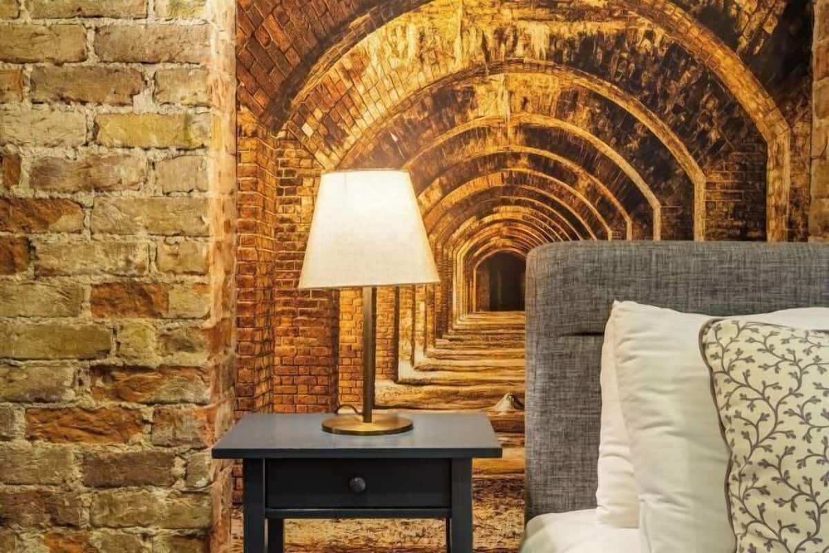 A room with a brick wall, a lamp on a nightstand, and a bed with a patterned pillow.