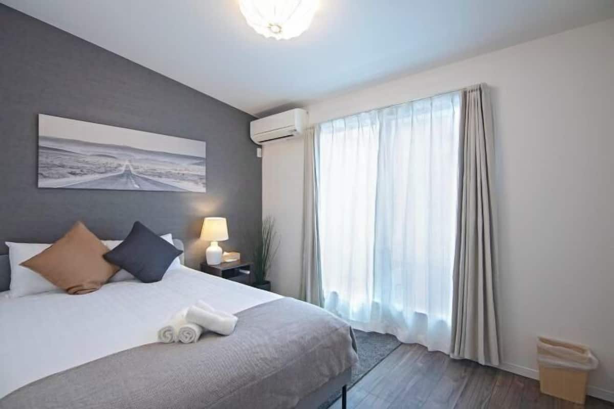 A hotel room with a bed, a nightstand, a lamp, an air conditioning unit, a window with curtains, and a wall-mounted artwork.