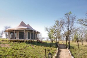 A single-story tent-style structure with a covered walkway leading to it, surrounded by a grassy area with sparse trees. - Kenzan Matawi Serengeti camp (Serengeti)