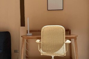 Huoneessa on puinen työpöytä, punottu tuoli ja valkoinen lamppu.