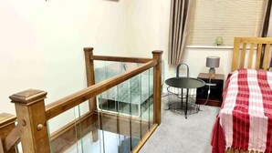 Una escalera con barandillas de madera y paneles de vidrio, una mesita redonda con una lámpara, una cama con colcha a cuadros y un ventanal con persianas.