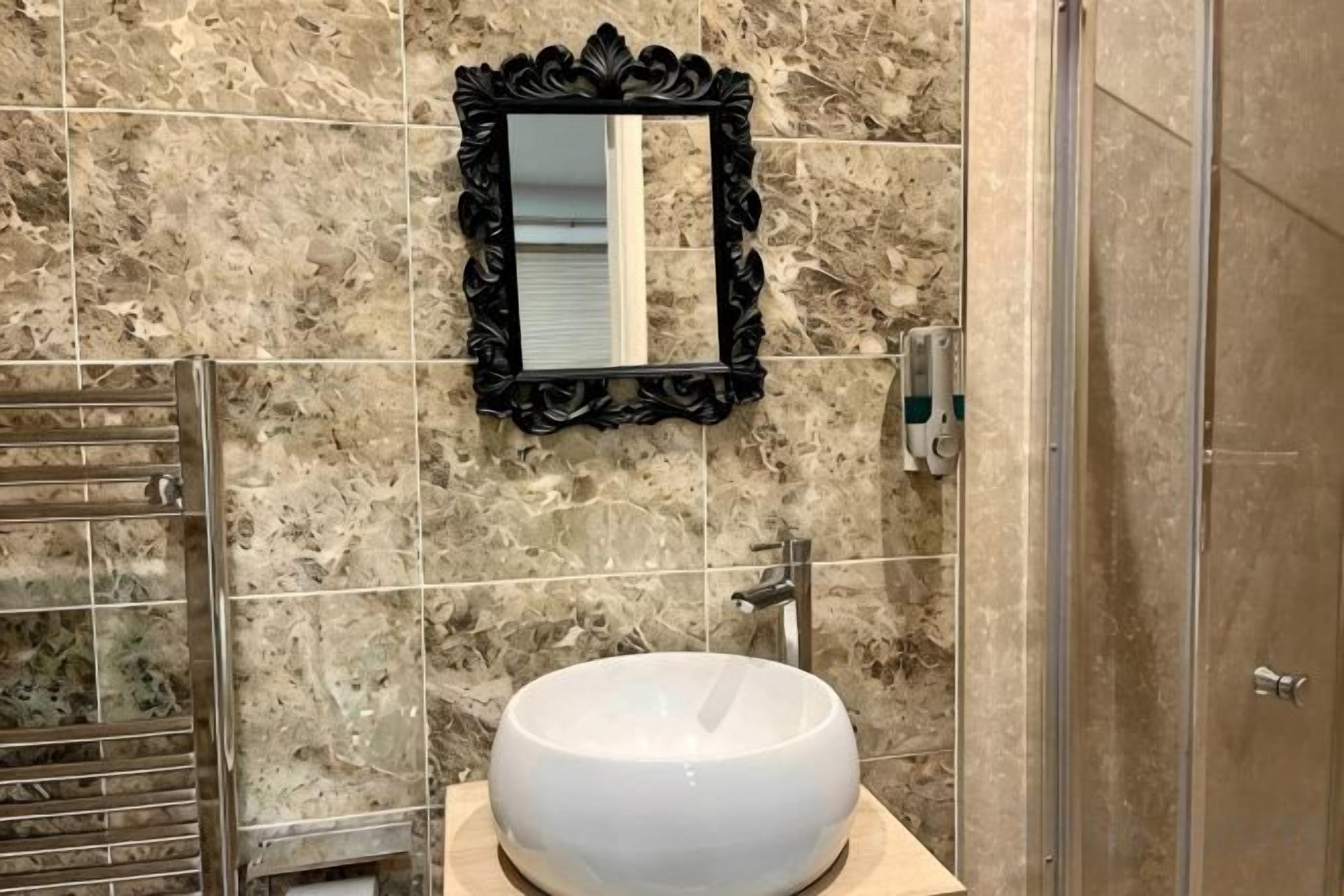 Un baño con pared de mármol, un lavamanos redondo, un espejo con marco negro y una puerta de ducha.