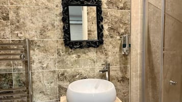 Un baño con pared de mármol, un lavamanos redondo, un espejo con marco negro y una puerta de ducha.