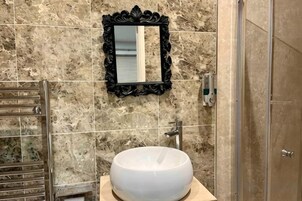 Un baño con pared de mármol, un lavamanos redondo, un espejo con marco negro y una puerta de ducha.