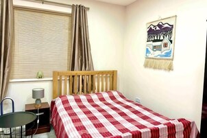 Un dormitorio con una cama individual, una silla, una mesita, una lámpara, una ventana con cortinas y un adorno en la pared.