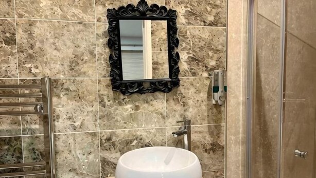 A bathroom with a marble wall, a round white sink, a black ornate mirror, and a glass shower enclosure. - AR-Rahman, 1 bed + 1 Living room apartment (Dartford)