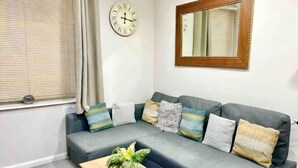 Una sala de estar con un sofá gris, una mesa de centro de madera con una planta, un reloj de pared y un espejo.