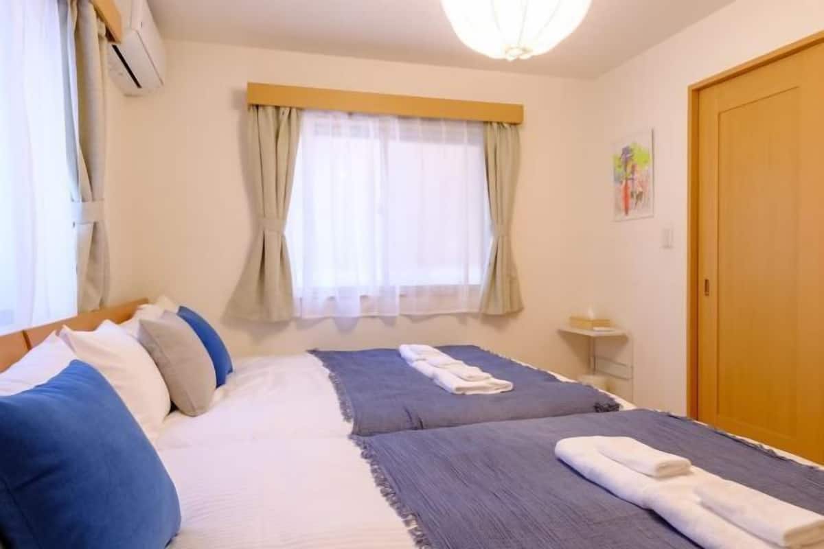 Una cama doble con edredón azul y blanco, una mesita de noche con lámpara y una ventana con cortinas.