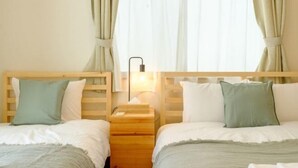 Ein Hotelzimmer mit zwei Betten, jeweils mit einem hölzernen Kopfteil, weißer Bettwäsche und grünen Kissen. Dazwischen befindet sich ein Nachttisch mit einer Lampe.