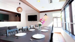 A dining area with a long table set for six, a flat-screen TV mounted on the wall, and a large window providing natural light. - GUEST HOUSE X (Tokyo)
