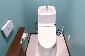 A toilet with a white seat and lid, a wall-mounted flush handle, and a toilet paper holder on the wall. - GUEST HOUSE X (Tokyo)