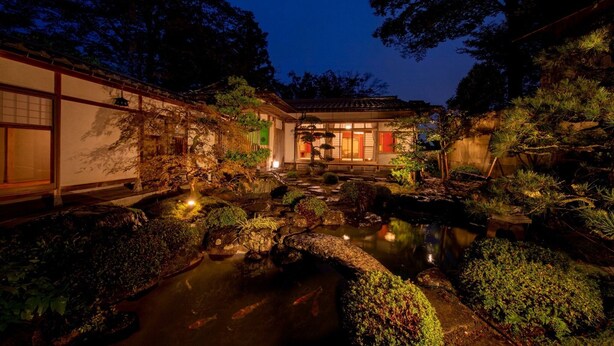 Een traditionele Japanse tuin in de schemering, met een vijver met koi karpers, rotsen en een houten gebouw met schuifdeuren.