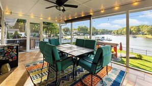 A covered patio with a table and chairs, large windows overlooking a lake, and a ceiling fan.