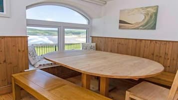 Une pièce dotée d’une grande table ovale en bois, de bancs, d’une vue dégagée sur l’extérieur grâce à une grande fenêtre en arc de cercle, et d’un tableau accroché au mur.