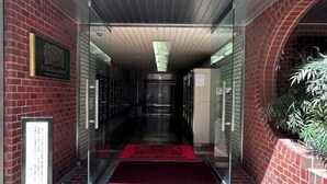 A hallway with a red carpet leading to a door, a sign on the wall, and a potted plant on the right. - Lions Mansion Dogenzaka SHIBUYA (Tokyo)