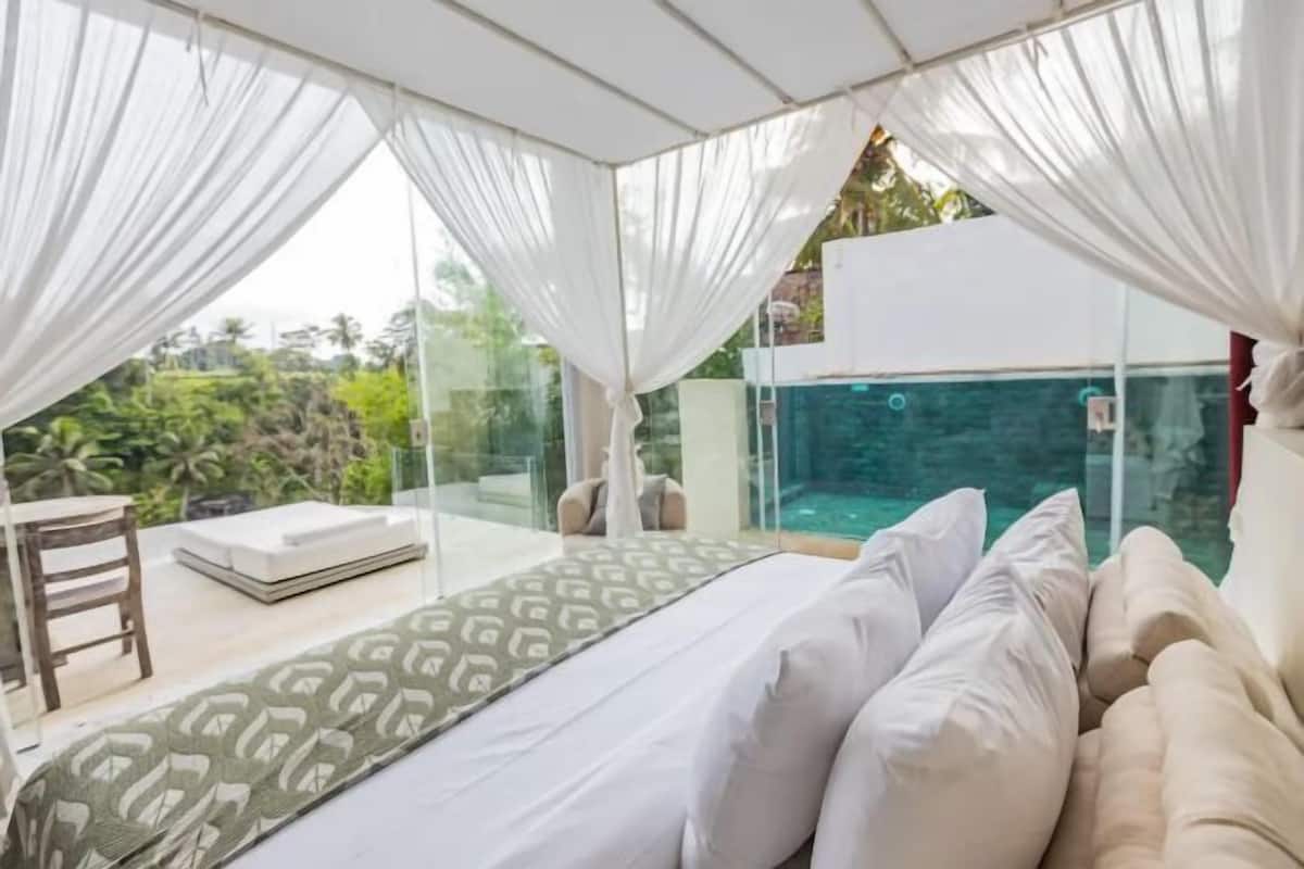 A room with a bed, a sofa, a table, and a glass wall overlooking a pool.