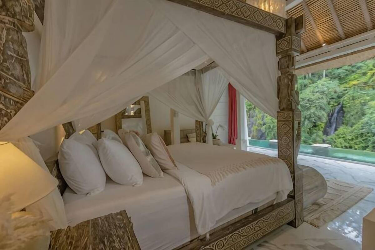A four-poster bed with white linens, a canopy, and a view of a lush garden.