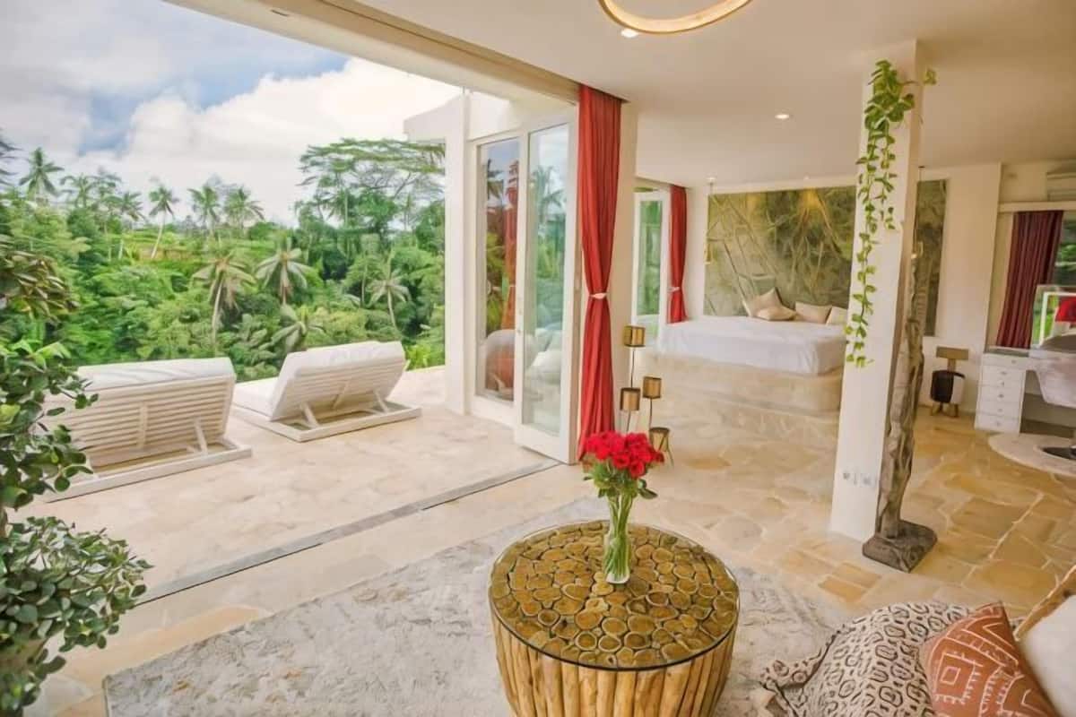 A modern hotel room with a large sliding glass door leading to a balcony, a round wooden table, and a bed with a view of the outdoors.