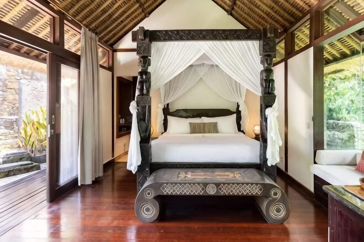A four-poster bed with a canopy, a wooden floor, and a view of greenery outside.