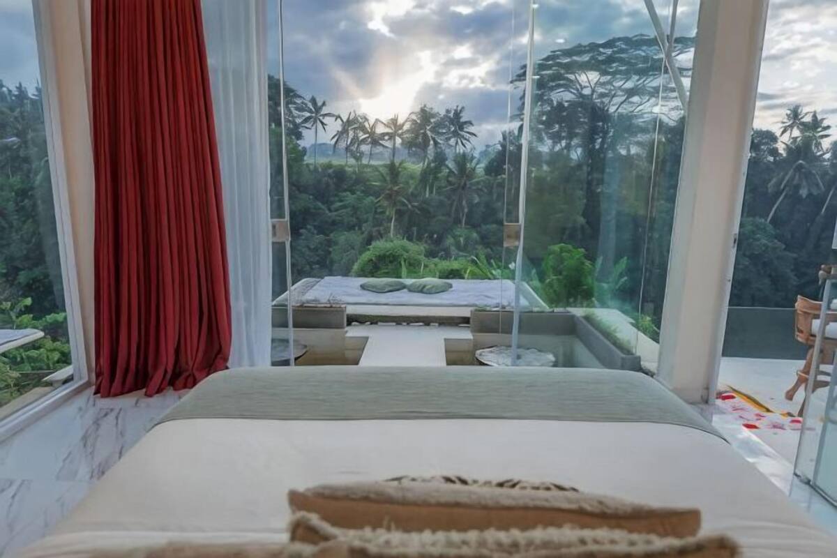 A bedroom with a large window, a bed, and a view of a balcony with a table and chairs.