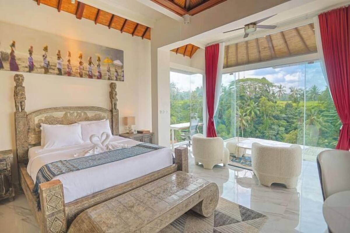 A bedroom with a large bed, a bench, a table, and a view of lush greenery.