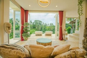 A spacious living room with a round coffee table, two sofas, and a view of lush greenery through large windows.