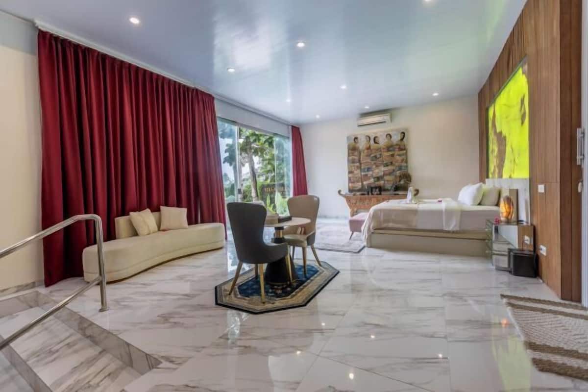 A modern hotel room with a marble floor, a sofa, a dining table with chairs, a bed, and a large window with red curtains.