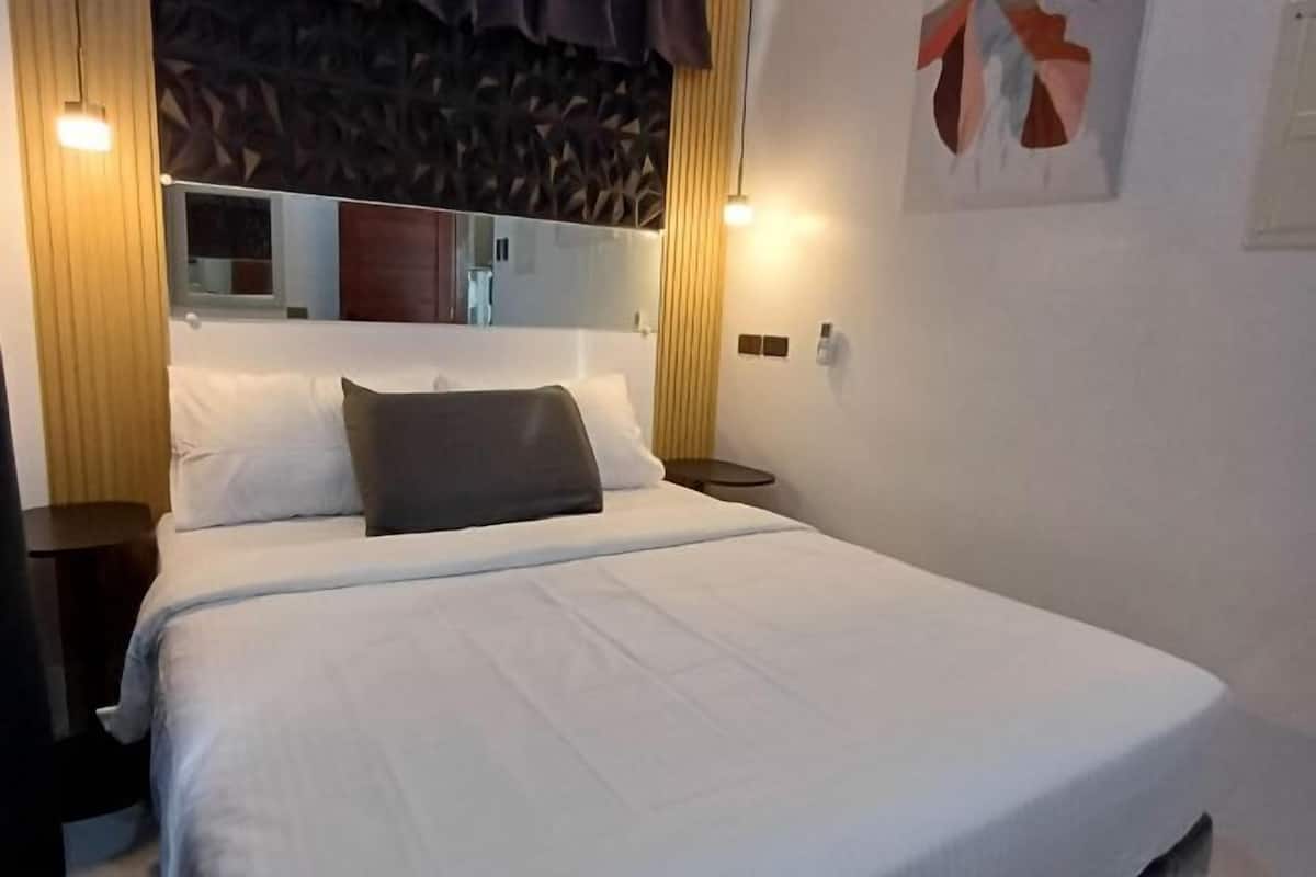 A neatly made bed with white linens and a single dark pillow, a bedside table with a lamp, and a wall-mounted mirror above the bed.