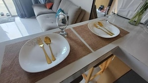 Et spisebord dækket med hvide tallerkener, guldbestik og et glas. Bordet står i et rum med en grå sofa og et vindue med gardiner.