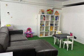 A room with a sectional sofa, a small table with chairs, a bookshelf, and a green carpet. - BelArt Golf Villa (Charneca de Caparica e Sobreda)