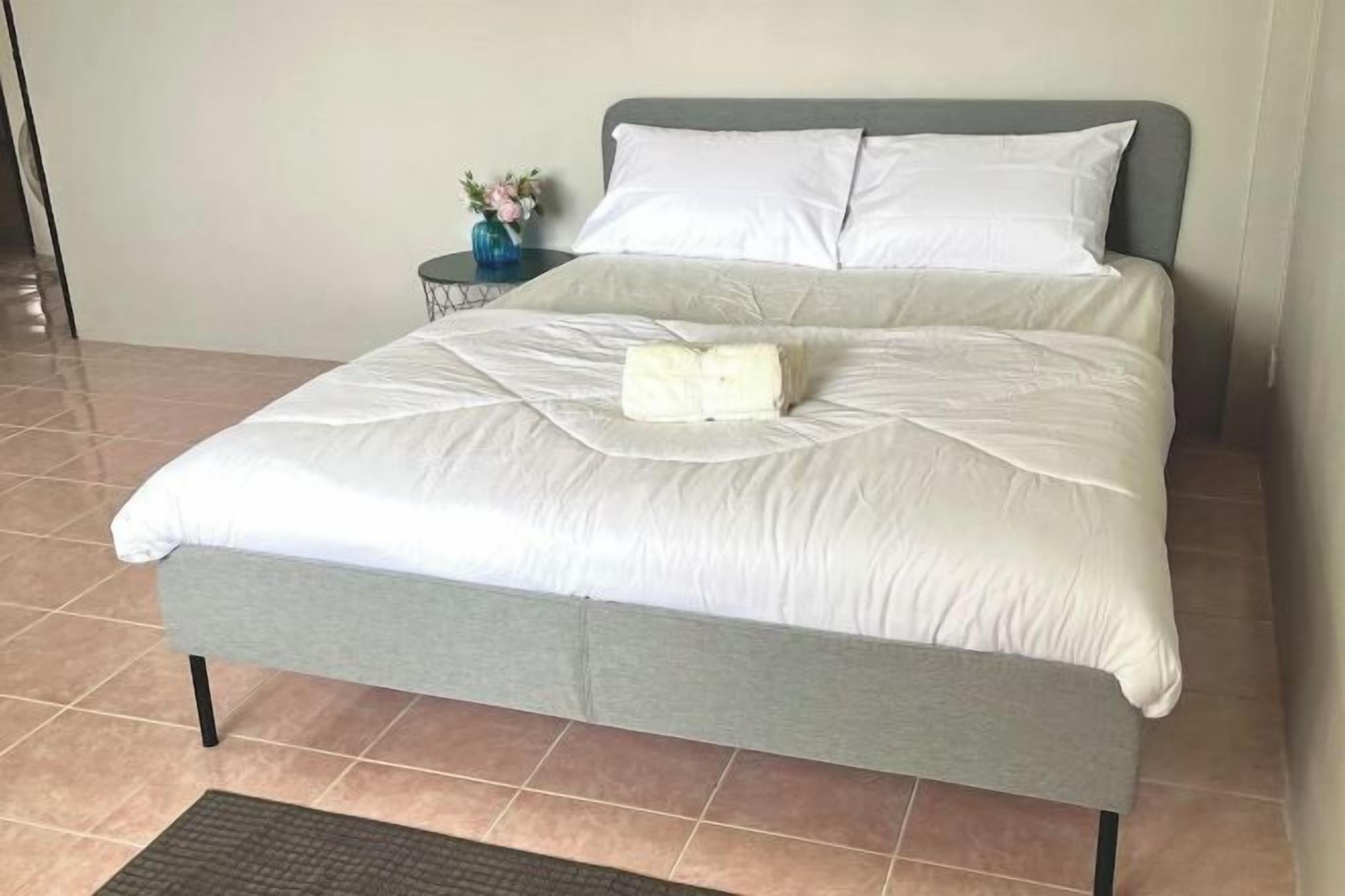 A neatly made bed with a grey headboard and white bedding, a small side table with a vase of flowers, and a tiled floor.