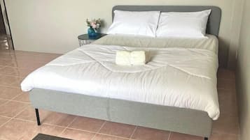 A neatly made bed with a grey headboard and white bedding, a small side table with a vase of flowers, and a tiled floor.