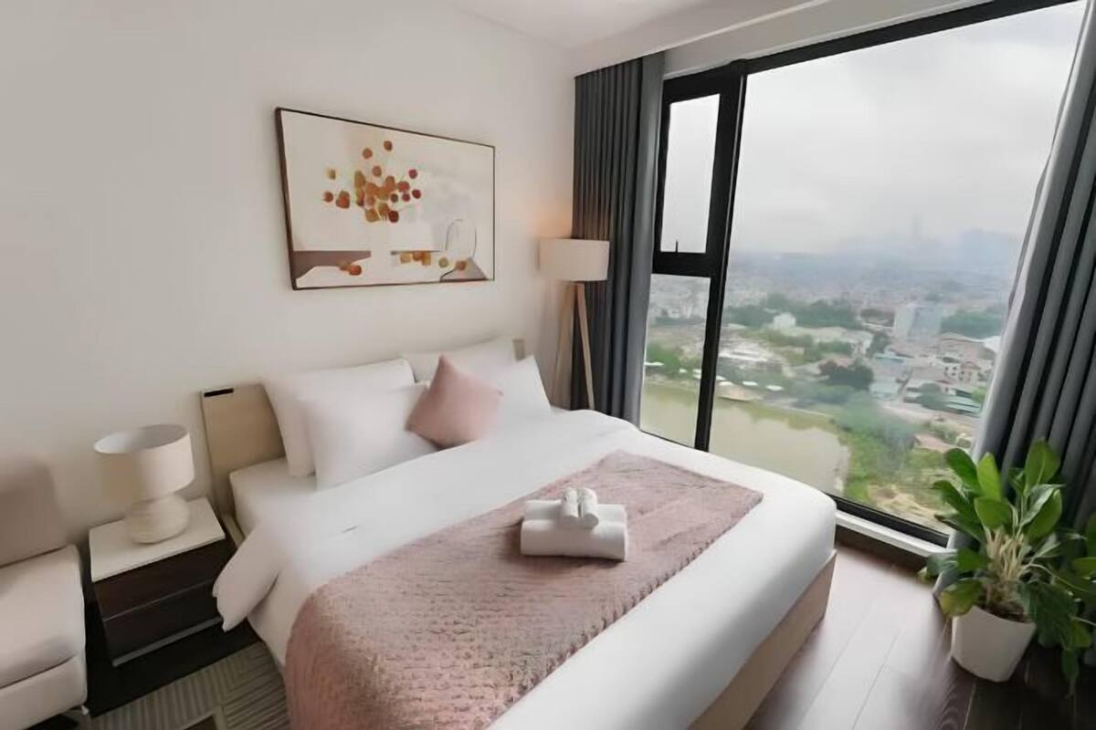 Quarto de hotel com uma cama grande, uma mesa de cabeceira, uma luminária, uma janela com vista para a cidade e um quadro na parede.