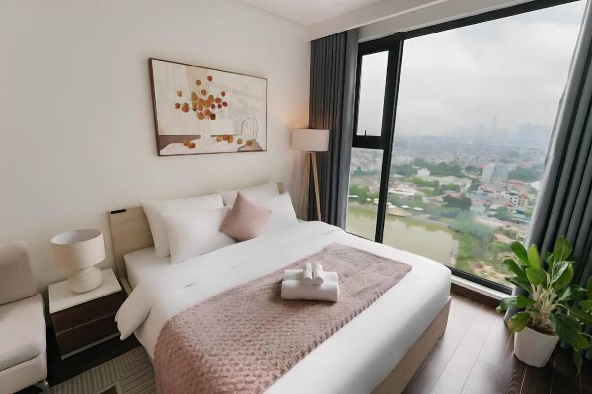 Quarto de hotel com uma janela grande, uma cama com manta rosa, mesa de cabeceira com abajur e um quadro na parede.