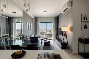 A modern hotel room with a dining area, a sofa, a standing lamp, and a large window with a view. - Fall-in-Love Suite - The Sun Avenue (Ho Chi Minh City)