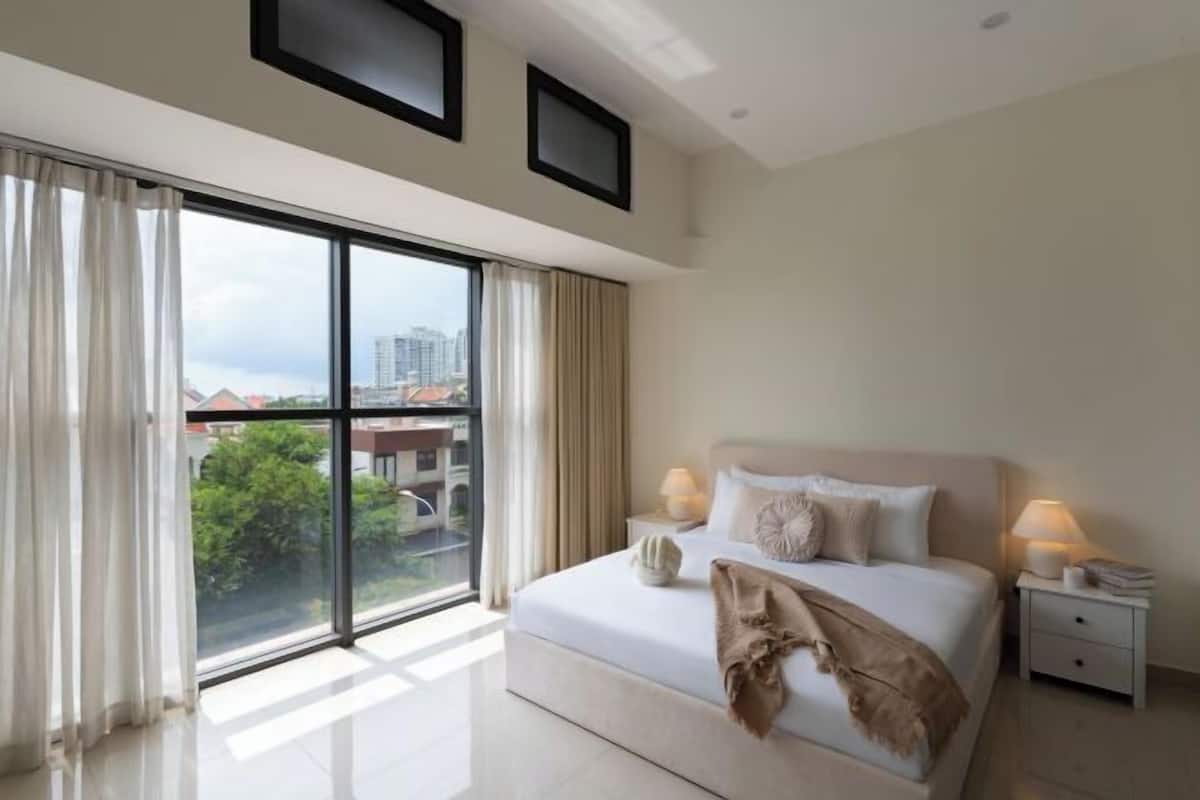 Une chambre d’hôtel moderne dotée d’un grand lit, de deux tables de chevet et offrant une vue sur la ville grâce à de grandes fenêtres du sol au plafond.